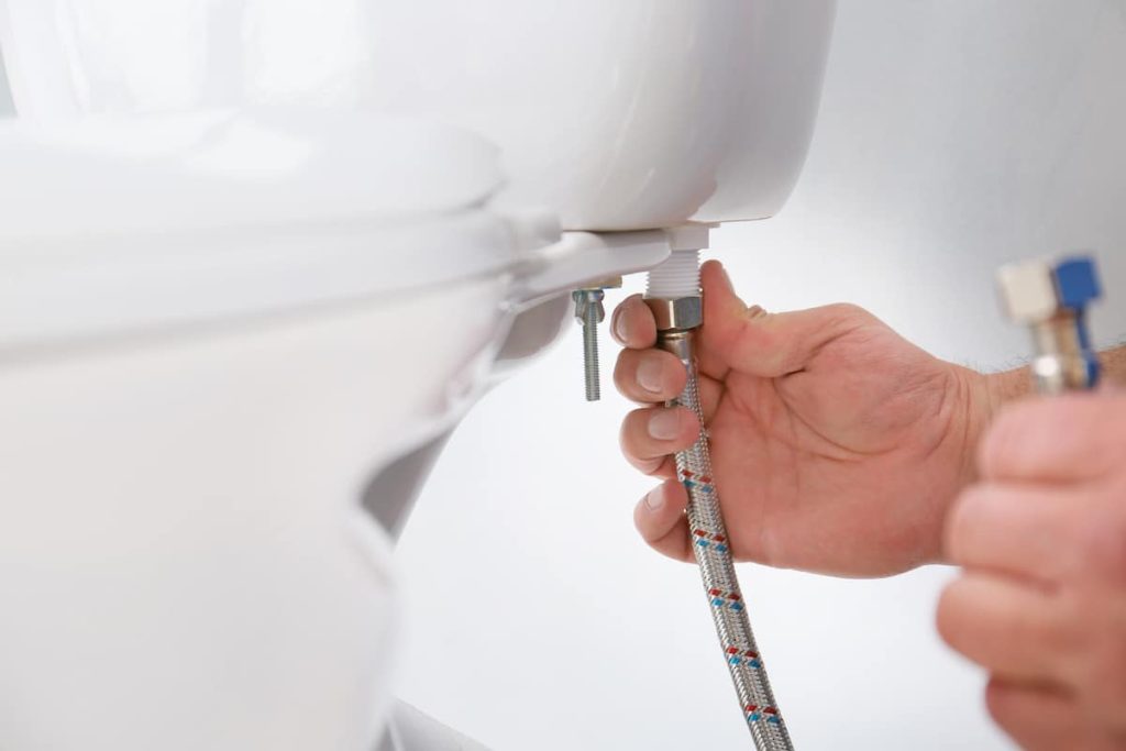 Close-up of a plumber connecting a flexible hose to the underside of a toilet cistern.