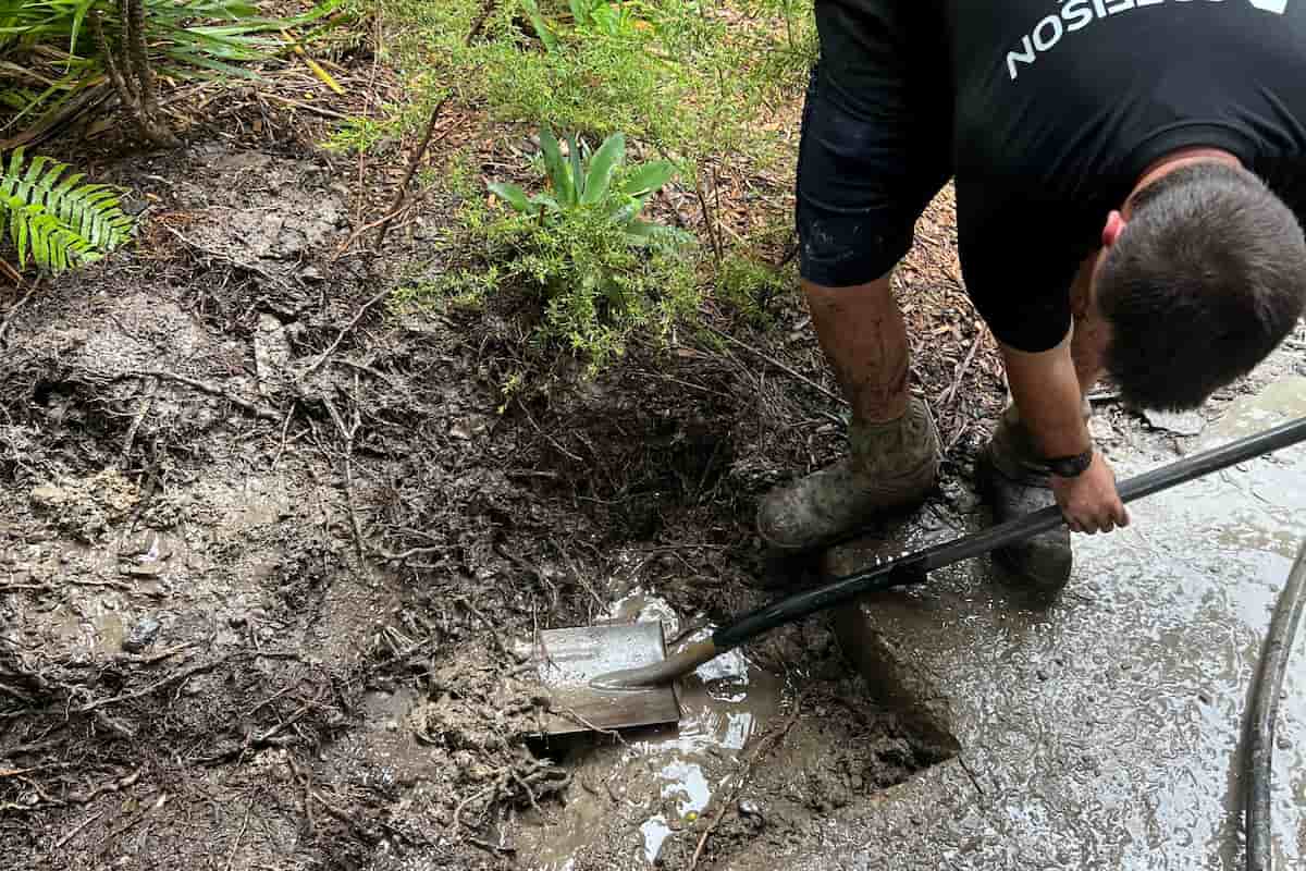 Coffison Plumbing technician digging to expose a burst pipe during a water leak excavation in a muddy garden area.